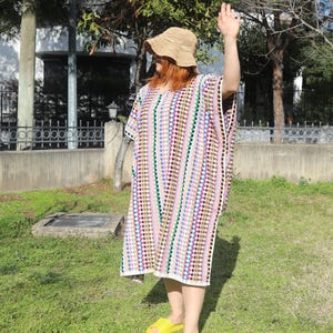 May include: A loose-fitting, knee-length crochet tunic featuring vertical stripes of multicoloured dots in shades of red, blue, green, and yellow. The garment has short sleeves and a round neckline. A tan crochet bucket hat is worn with the tunic.