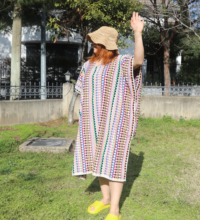 May include: A loose-fitting, knee-length crochet tunic featuring vertical stripes of multicoloured dots in shades of red, blue, green, and yellow. The garment has short sleeves and a round neckline. A tan crochet bucket hat is worn with the tunic.
