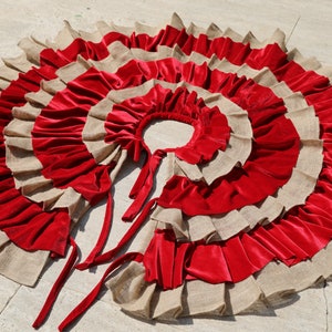May include: A red and beige burlap Christmas tree skirt with multiple layers of ruffles. The skirt has a center opening and ties at the back.