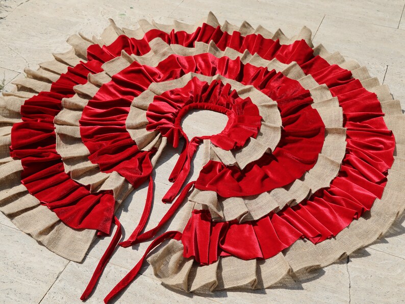 May include: A red and beige burlap Christmas tree skirt with multiple layers of ruffles. The skirt has a center opening and ties at the back.