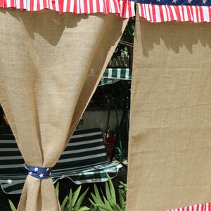 May include: Two burlap curtains with red, white, and blue ruffled trim featuring stars and stripes. The curtains are hung on a rod and are partially open, revealing a green and white striped chair in the background.