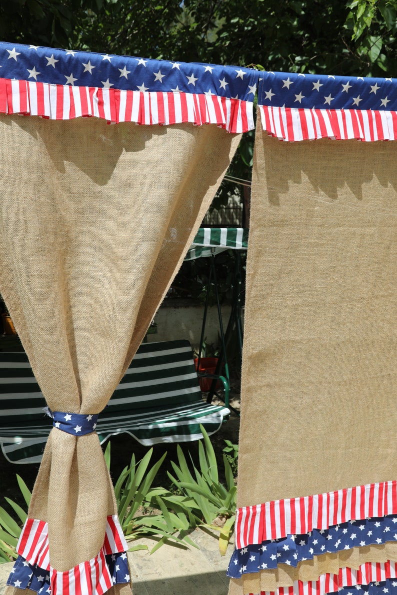 May include: Two burlap curtains with red, white, and blue ruffled trim featuring stars and stripes. The curtains are hung on a rod and are partially open, revealing a green and white striped chair in the background.