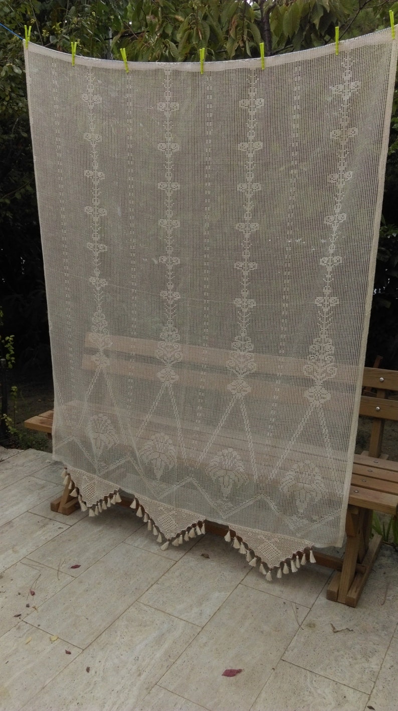 May include: A white crocheted curtain with a delicate floral pattern and fringe trim. The curtain is hanging from a wooden bench.