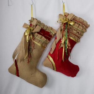 May include: Two Christmas stockings, one is red with gold trim and the other is burlap with gold and green trim. Both stockings have a gold bow and are decorated with beads and ribbons.