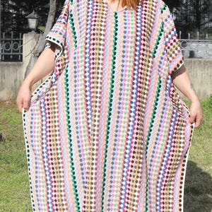 May include: A crochet kaftan featuring a vertical pattern of multicoloured dots in shades of pink, blue, green, yellow, and black, paired with a tan crochet sun hat and bright yellow slip-on sandals. The garment has a V-neckline and short sleeves.
