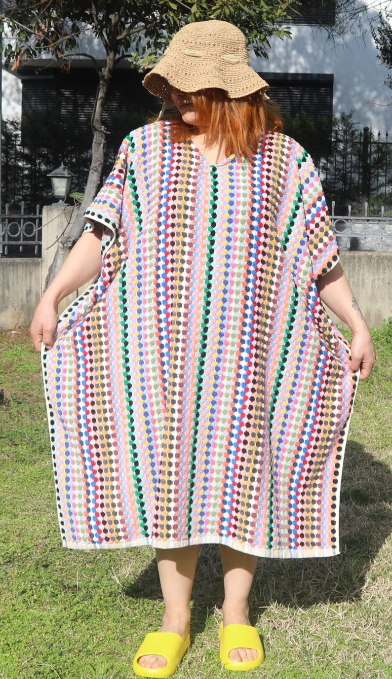 May include: A crochet kaftan featuring a vertical pattern of multicoloured dots in shades of pink, blue, green, yellow, and black, paired with a tan crochet sun hat and bright yellow slip-on sandals. The garment has a V-neckline and short sleeves.