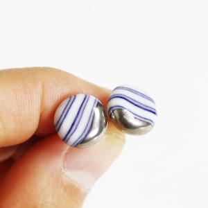 May include: A pair of round stud earrings. Each earring features a white base with blue stripes and a silver-colored metal backing. The earrings are held between fingers against a white background.