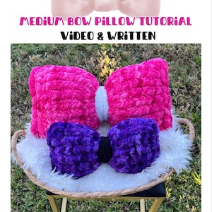 May include: A handmade bow-shaped pillow in bright pink and purple, resting on a white fluffy surface within a woven tray. The pillow has a black center and is surrounded by faux fur. Text reads "MEDIUM BOW PILLOW TUTORIAL VIDEO & WRITTEN".