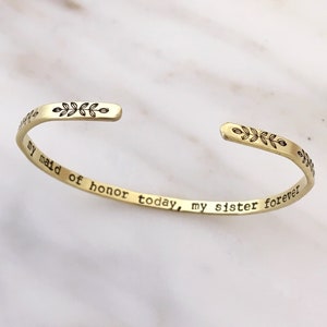 Golden brass skinny stacking cuff bracelet features a simple, hand stamped leaf design across the front with the saying my maid of honor today, my sister forever stamped inside. Text can be personalized. Outside finish can be matte or polished.