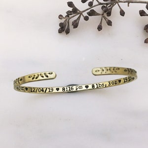 May include: A gold-colored cuff bracelet with engraved text and floral accents. The text includes a date, time, and weight: 8 lbs, 3 oz. The bracelet is open-ended and rests on a white surface.
