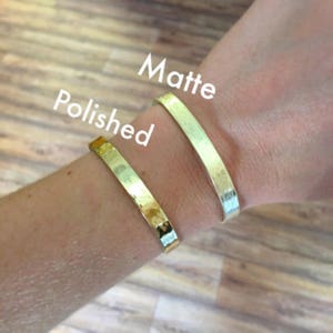 May include: Two gold-colored bangles, one matte and one polished, are displayed on a wrist. The text "Polished" and "Matte" are written above the bangles. The bangles are made of a hammered metal.