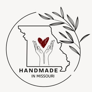 K&ouml;nnte beinhalten: Rundes Logo mit schwarzer Umrisslinie von Missouri, rotem Herz in H&auml;nden und floralem Element. Der Text "HANDMADE IN MISSOURI" steht darunter. Das Design ist in Schwarz und Rot auf cremefarbenem Hintergrund.
