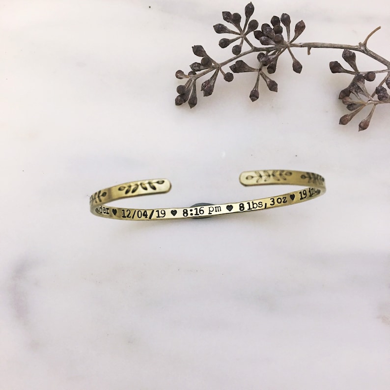 May include: A gold-colored cuff bracelet with engraved text and floral accents. The text includes a date, time, and weight: 8 lbs, 3 oz. The bracelet is open-ended and rests on a white surface.
