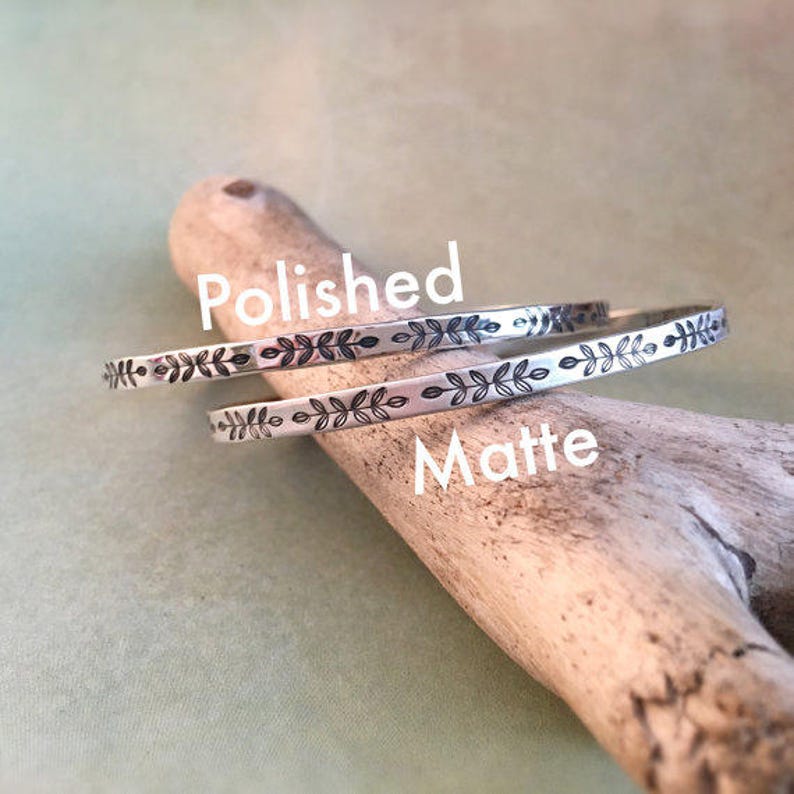 May include: Two silver bangles with leaf patterns, one polished and one matte, are displayed on a piece of driftwood. The word "Polished" is above the top bangle, and "Matte" is above the bottom one.