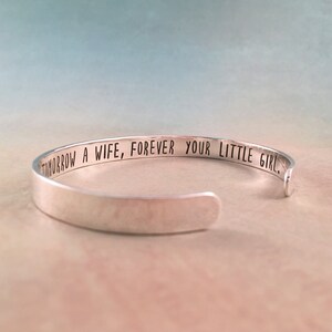 May include: A silver cuff bracelet with the engraved text: "TOMORROW A WIFE, FOREVER YOUR LITTLE GIRL." The bracelet is open-ended and has a smooth, polished finish. The background is a soft gradient of blue and peach.