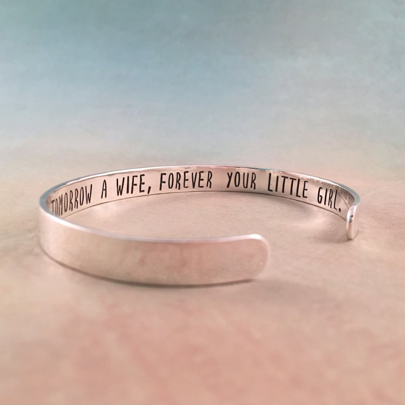 May include: A silver cuff bracelet with the engraved text: "TOMORROW A WIFE, FOREVER YOUR LITTLE GIRL." The bracelet is open-ended and has a smooth, polished finish. The background is a soft gradient of blue and peach.
