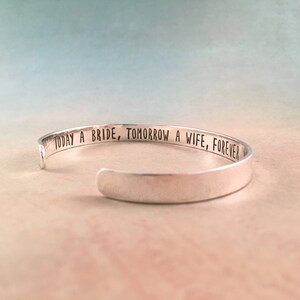 May include: A silver cuff bracelet with the engraved text "TODAY A BRIDE, TOMORROW A WIFE, FOREVER". The open-ended bracelet is displayed on a soft, neutral background with a gradient of blue and peach tones.