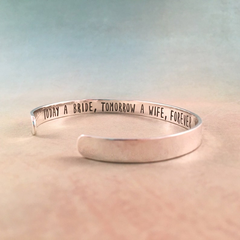 May include: A silver cuff bracelet with the engraved text "TODAY A BRIDE, TOMORROW A WIFE, FOREVER". The open-ended bracelet is displayed on a soft, neutral background with a gradient of blue and peach tones.