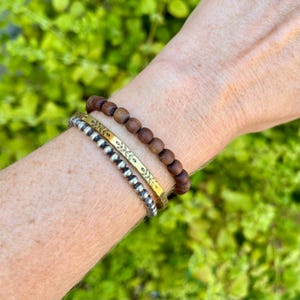 A gold brass cuff bracelet with a symmetric floral design on the surface. stacked with a silver beaded bracelet and a wooden beaded bracelet.