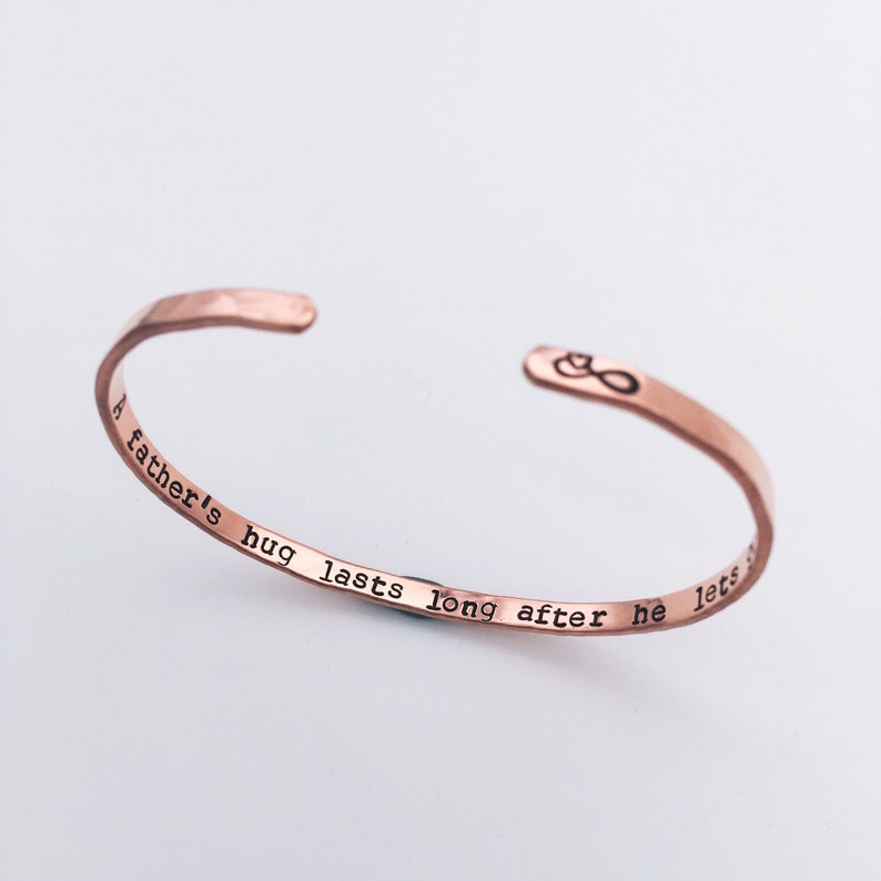 Thin stacking cuff bracelet in rose toned copper. Hand stamped inside is A fathers hug lasts long after he lets go with an infinity sign and heart design on the outside right end of the cuff. Simple, minimalist design to remember a lost loved one