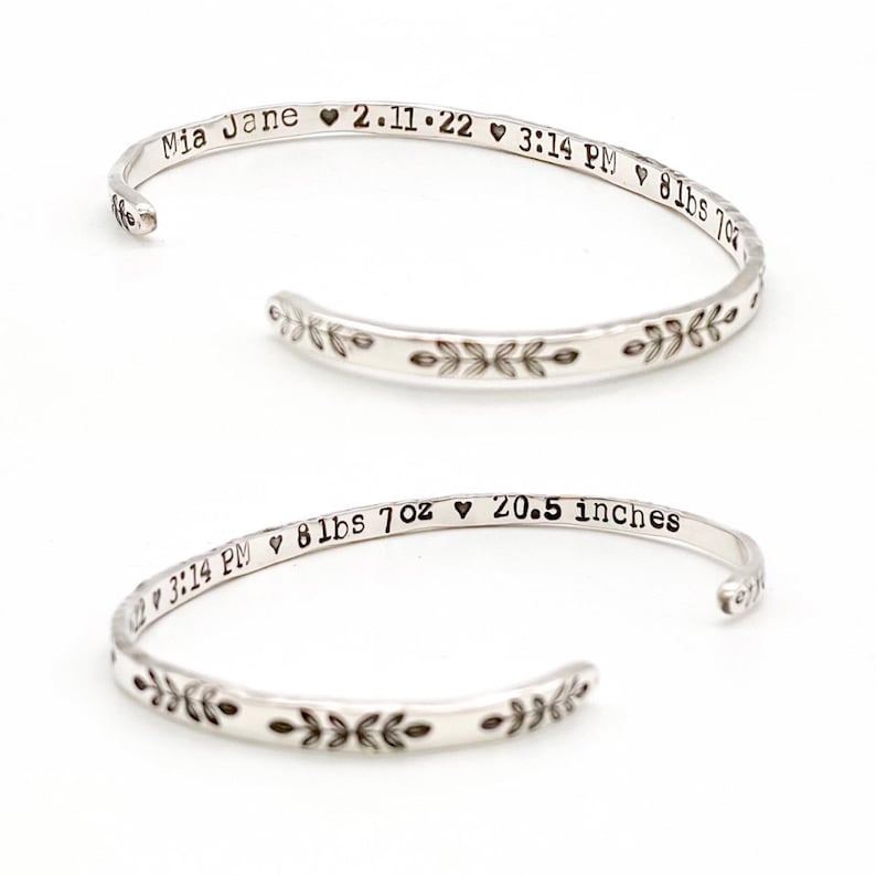 May include: Two silver-colored, open-ended bracelets. The bracelets have a floral design and stamped text. One bracelet reads "Mia Jane 2.11.22 3:14 PM 8lbs 7oz 20.5 inches". The other bracelet has similar text.