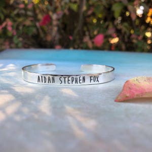 one fourth inch wide sterling silver cuff bracelet customized with baby's name on front, in an uppercase handwritten font. Birth stats are stamped inside the bracelet.