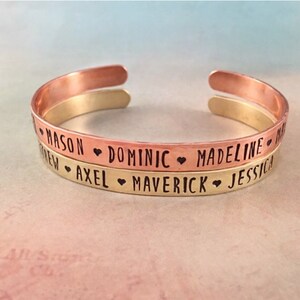 May include: Two stacked copper and gold bracelets with engraved names: Mason, Dominic, Madeline, Axel, Maverick, and Jessica. Each name is separated by a heart symbol.