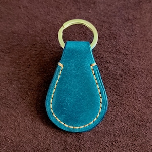 May include: Teal leather teardrop-shaped keychain with a gold-colored key ring. The edges are stitched with light brown thread. The keychain is on a brown background.