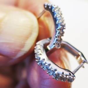 May include: A pair of silver hoop earrings, encrusted with small, sparkling gemstones. The earrings are held up, showcasing their curved design and the way the light catches the stones. A close-up shot highlights the jewelry's details.