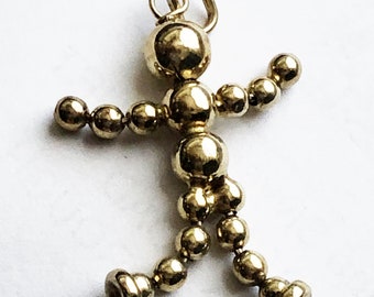 May include: A gold-coloured charm in the shape of a stick figure. The charm is made of small, round beads connected to form the body, arms, and legs. It has a loop at the top for attaching to a necklace or bracelet.