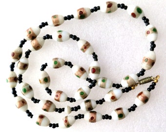 May include: A white oval bead necklace with gold, red and green accents. The beads are strung on a black cord.