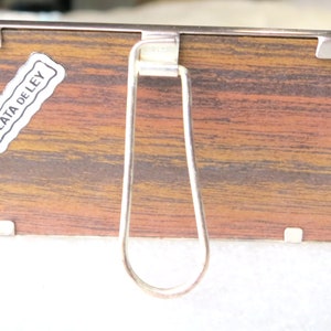 May include: A silver-coloured picture frame with a wood-grain backing. The frame has a metal stand on the back and a label that reads "PLATA DE LEY".