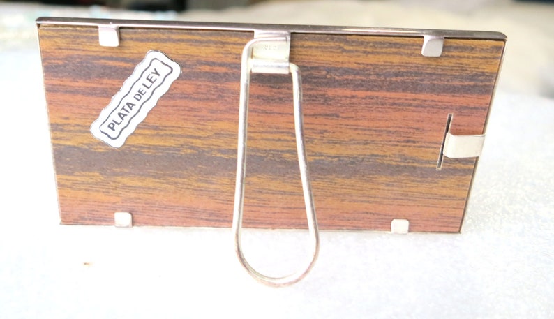 May include: A silver-coloured picture frame with a wood-grain backing. The frame has a metal stand on the back and a label that reads "PLATA DE LEY".