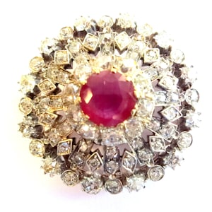 Puede incluir: Un broche redondo con un gran rubí rojo rodeado por un halo de diamantes más pequeños.