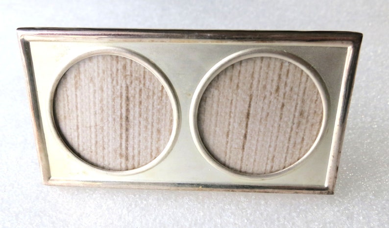 May include: A silver-toned double picture frame with two round openings. The frame has a textured surface and a simple design.