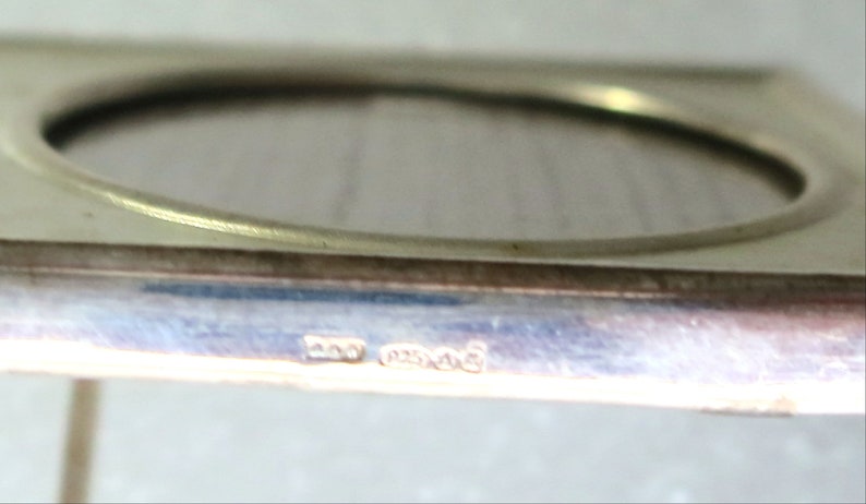 May include: A silver-toned square picture frame with a rounded opening. The frame has a hallmark on the bottom edge that reads "925 Mexico".