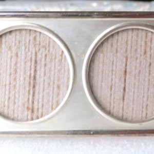 May include: A silver-toned double picture frame with two round openings. The frame has a smooth, polished finish and a simple, elegant design.