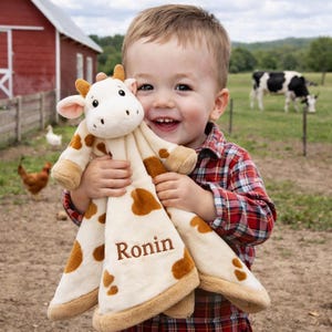 May include: A plush giraffe security blanket with brown spots and trim is held by a smiling child. The blanket has the name "Ronin" embroidered on it. The background includes a red barn, chickens, and a cow in a field.