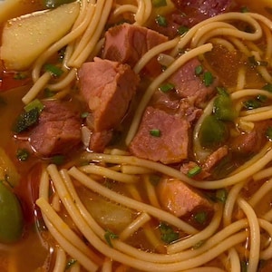 May include: A bowl of soup with spaghetti noodles, chunks of meat, and vegetables. The broth is a reddish-orange color, and the bowl has a floral pattern around the rim. The soup appears to be a hearty and flavorful meal.