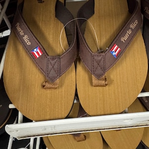 May include: A pair of brown and tan flip-flops with the words "Puerto Rico" printed on the straps. The straps also feature the Puerto Rican flag. The flip-flops are displayed on a white shelf.