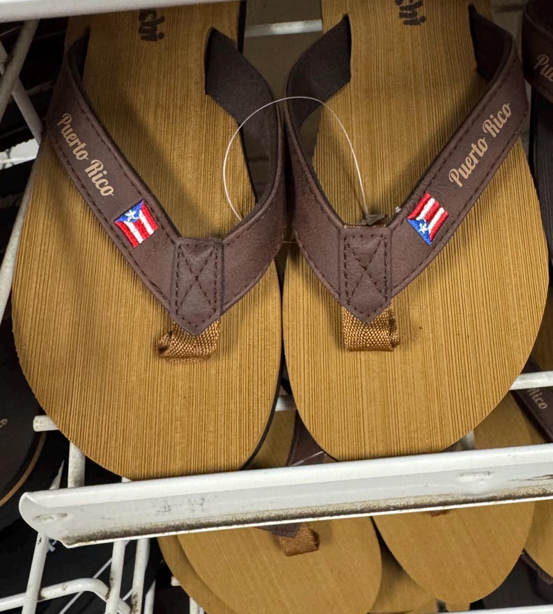 May include: A pair of brown and tan flip-flops with the words "Puerto Rico" printed on the straps. The straps also feature the Puerto Rican flag. The flip-flops are displayed on a white shelf.