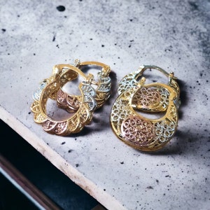 May include: Two pairs of gold hoop earrings with intricate filigree designs. The earrings are made of three different shades of gold, creating a unique and elegant look.