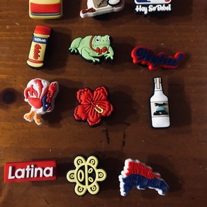 Puede incluir: Una colecci&oacute;n de 11 dijes de zapatos coloridos con temas latinoamericanos. Los dijes incluyen un logotipo de b&eacute;isbol rojo, blanco y azul con el texto "Hay So Bibel", una rana verde con una corbata de mo&ntilde;o roja, una flor de hibisco roja, un sol amarillo con una cara, un gallo rojo y blanco, una lata roja y blanca con el texto "Cafe Bustelo", una botella roja y blanca con el texto "Goya Adobo", una botella blanca con una etiqueta azul, un dije rojo y blanco con el texto "Ole!", un dije rojo y blanco con el texto "Latina", y un dije azul, rojo y blanco con el texto "Rep&uacute;blica Dominicana".