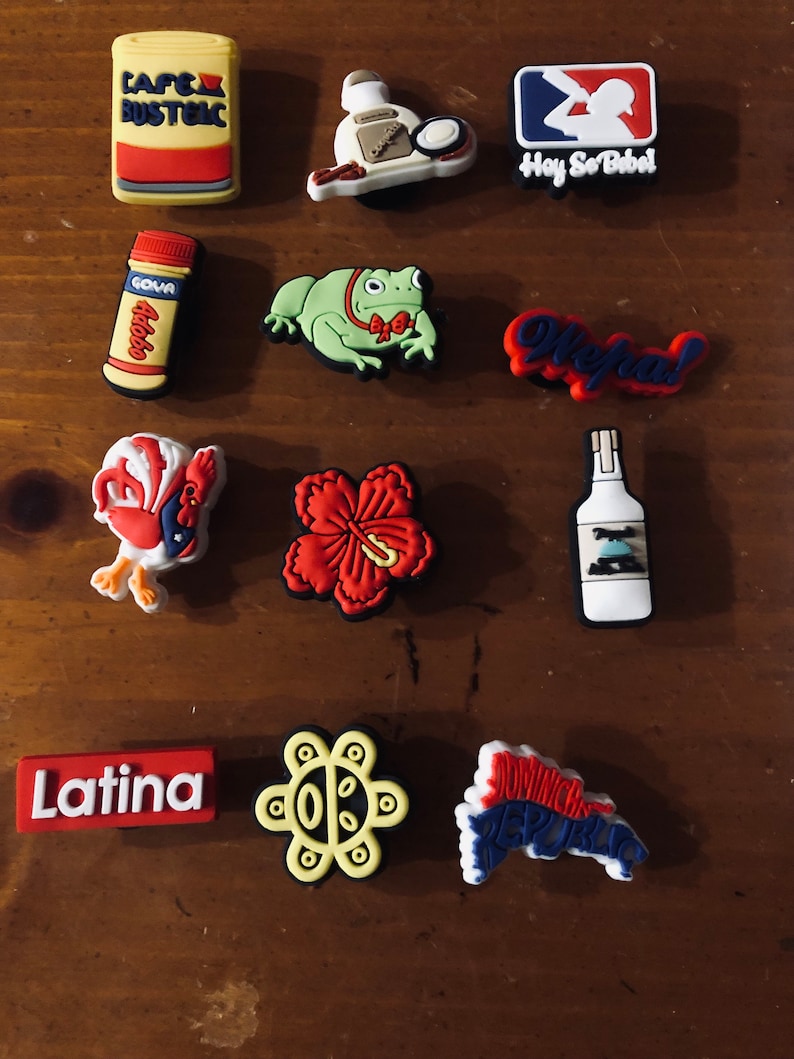 Puede incluir: Una colecci&oacute;n de 11 dijes de zapatos coloridos con temas latinoamericanos. Los dijes incluyen un logotipo de b&eacute;isbol rojo, blanco y azul con el texto "Hay So Bibel", una rana verde con una corbata de mo&ntilde;o roja, una flor de hibisco roja, un sol amarillo con una cara, un gallo rojo y blanco, una lata roja y blanca con el texto "Cafe Bustelo", una botella roja y blanca con el texto "Goya Adobo", una botella blanca con una etiqueta azul, un dije rojo y blanco con el texto "Ole!", un dije rojo y blanco con el texto "Latina", y un dije azul, rojo y blanco con el texto "Rep&uacute;blica Dominicana".