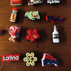 Puede incluir: Una colecci&oacute;n de 12 dijes de zapatos coloridos con varios temas latinoamericanos. Los dijes incluyen una rana, una flor, un gallo, una botella de licor, una lata de caf&eacute;, una botella de jugo de guayaba, un logotipo de b&eacute;isbol, una flor, un mapa de la Rep&uacute;blica Dominicana, un texto Latina, un s&iacute;mbolo de la Rep&uacute;blica Dominicana y un texto que dice "Hey So Babel".