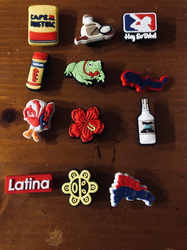 Puede incluir: Una colecci&oacute;n de 12 dijes de zapatos coloridos con varios temas latinoamericanos. Los dijes incluyen una rana, una flor, un gallo, una botella de licor, una lata de caf&eacute;, una botella de jugo de guayaba, un logotipo de b&eacute;isbol, una flor, un mapa de la Rep&uacute;blica Dominicana, un texto Latina, un s&iacute;mbolo de la Rep&uacute;blica Dominicana y un texto que dice "Hey So Babel".