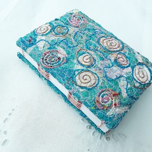 May include: A turquoise blue fabric-covered journal with a white spine and decorative stitching in the shape of swirls. The cover is made of a variety of fabrics and textures, creating a unique and handmade look.