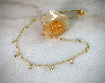 May include: A gold chain necklace with small clear stones dangling from it. The necklace is laying on a white surface with a large orange crystal.