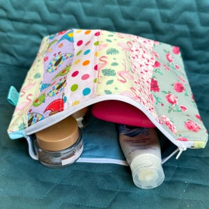 May include: A patchwork cosmetic bag with various colourful patterns, including flamingos, polka dots, and floral designs. The bag is open, revealing several cosmetic products inside. The bag has a white zip and a small blue tag.