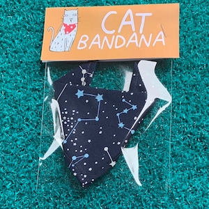 May include: A blue and white cat bandana with a constellation pattern. The bandana is packaged in a clear plastic bag with a yellow label that says "CAT BANDANA" and features a cartoon cat wearing a red bandana.